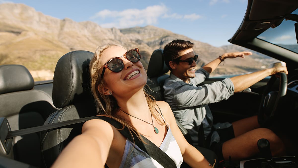 Pareja sonriente con gafas de sol tomando un selfie en un coche descapotable con montañas de fondo.
